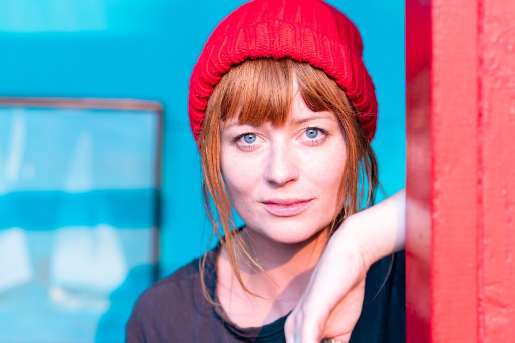 Cute ginger girl with blue eyes and red beanie on a blue wall